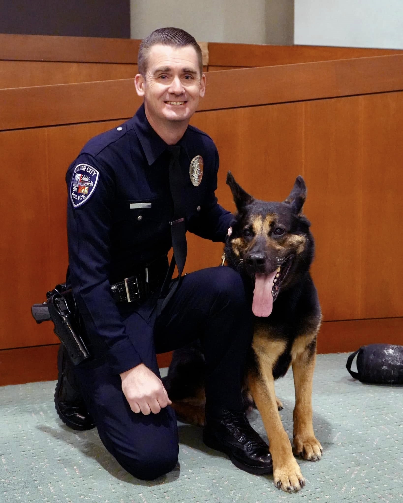 K9 Officer kneels down next to his four-legged K9 partner Ander in Council Chambers during his retirement ceremony